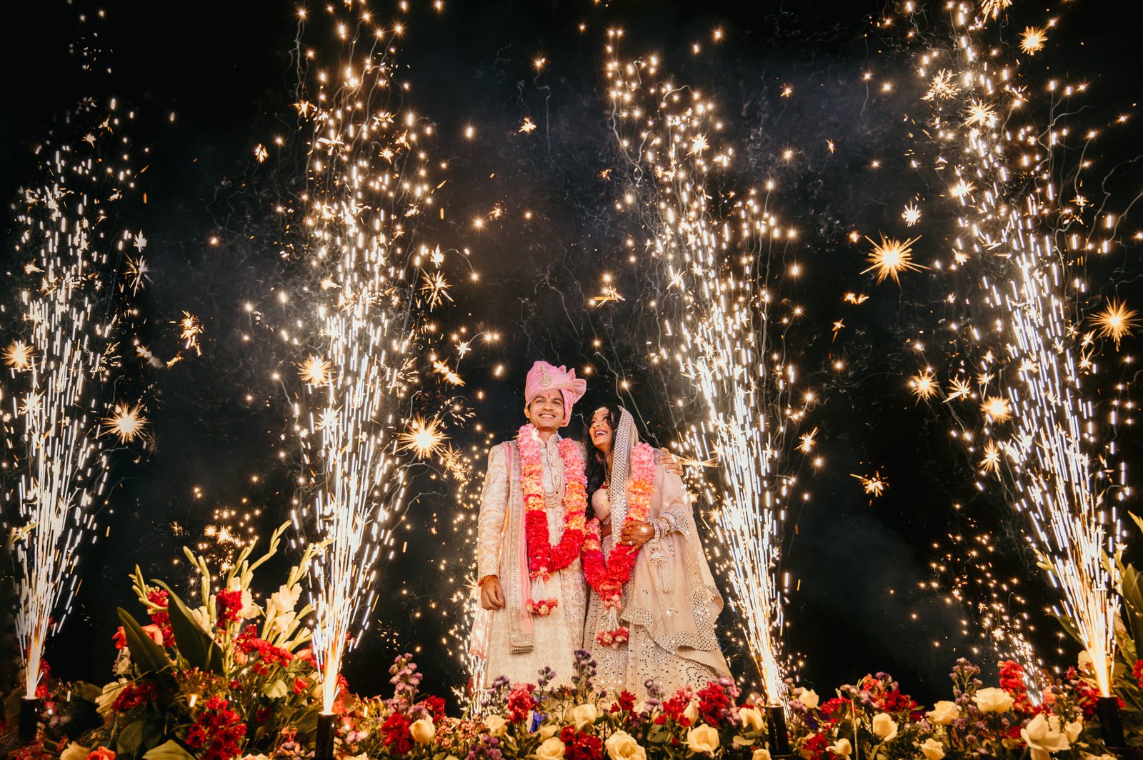 A newly married couple stands together surrounded by a shower of bright fireworks and red flowers.