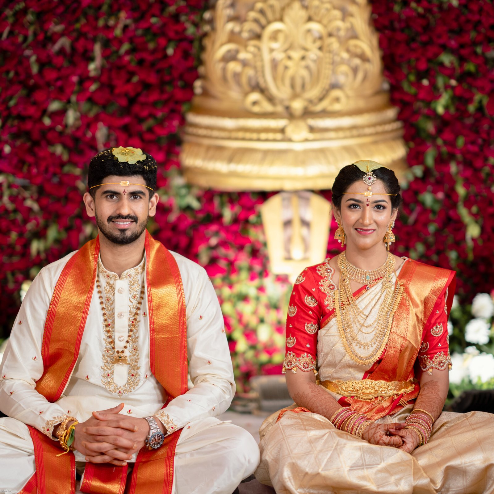 South Indian bride and groom sitting together in traditional wedding attire against a red floral backdrop