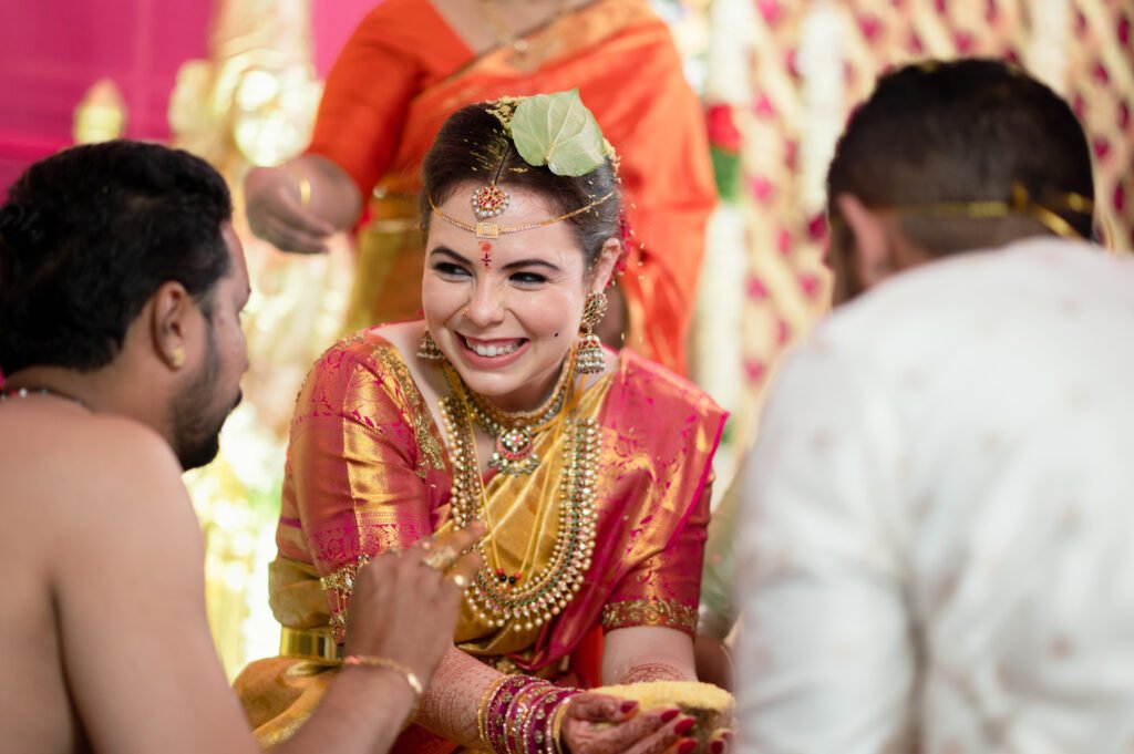 Bride laughing during a ritual, seated between family members with hands adorned in mehendi and jewelry, wearing a pink and gold saree
