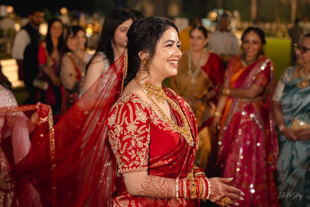 Bride in a red and gold lehenga with a sheer dupatta, smiling candidly amid guests during an evening wedding celebration