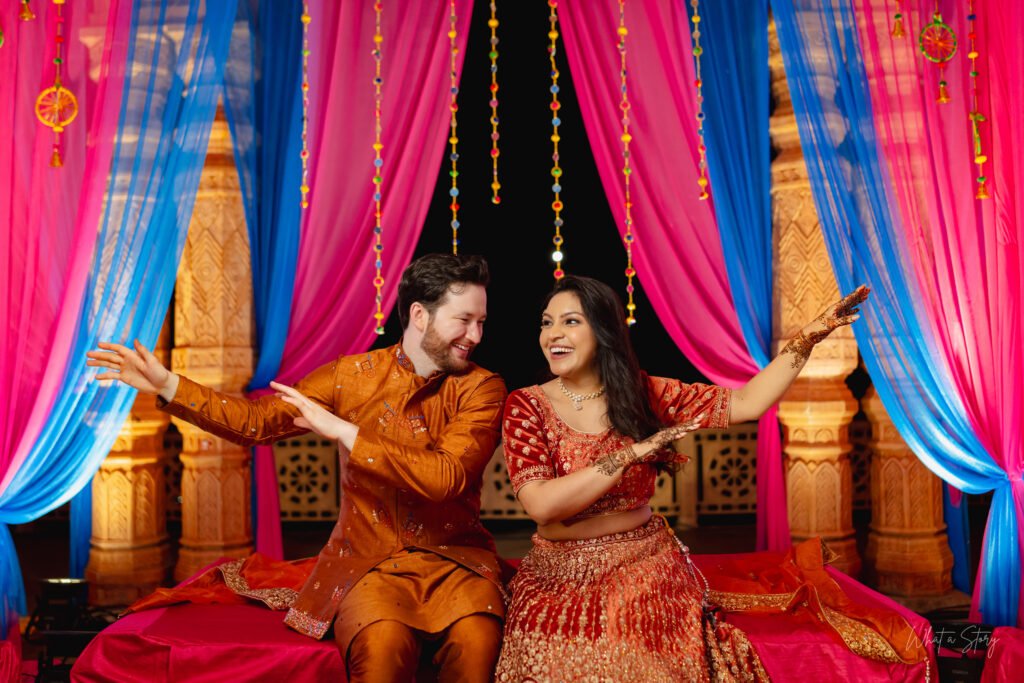 Couple dancing playfully on a decorated stage with pink and blue drapes during their sangeet celebration