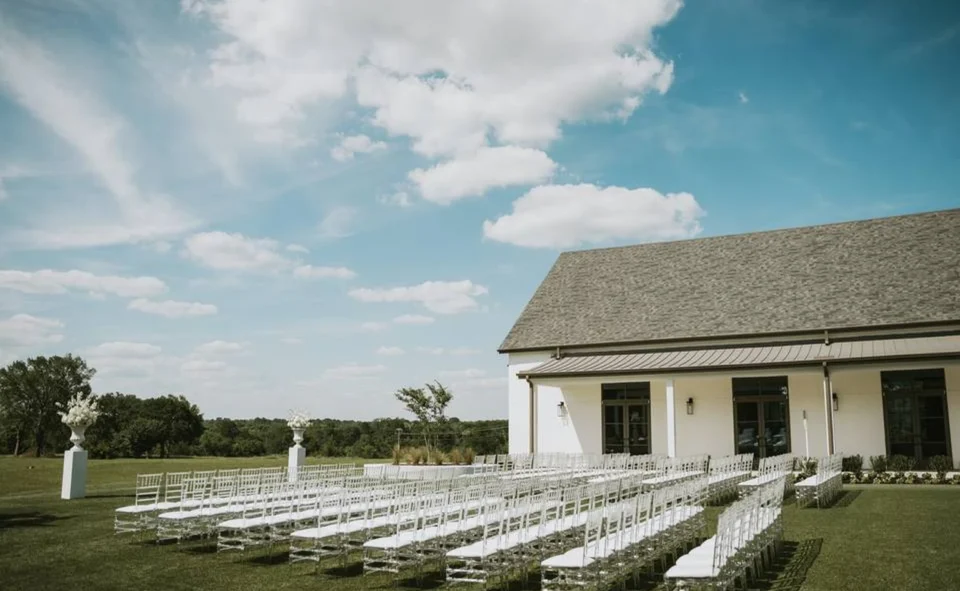 Outdoor wedding ceremony venue with rows of white chiavari chairs on a lawn beside a white farmhouse in montgomery, Houston.