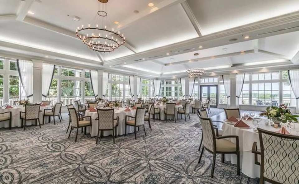 Elegant event ballroom with crystal chandeliers, floor-to-ceiling windows, patterned carpet, and round tables set with white linens for a formal dinner.