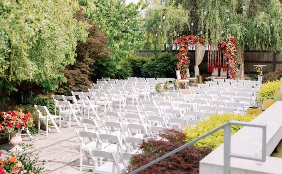 Outdoor wedding ceremony venue with rows of white folding chairs facing a floral arch, string lights, and lush garden greenery.