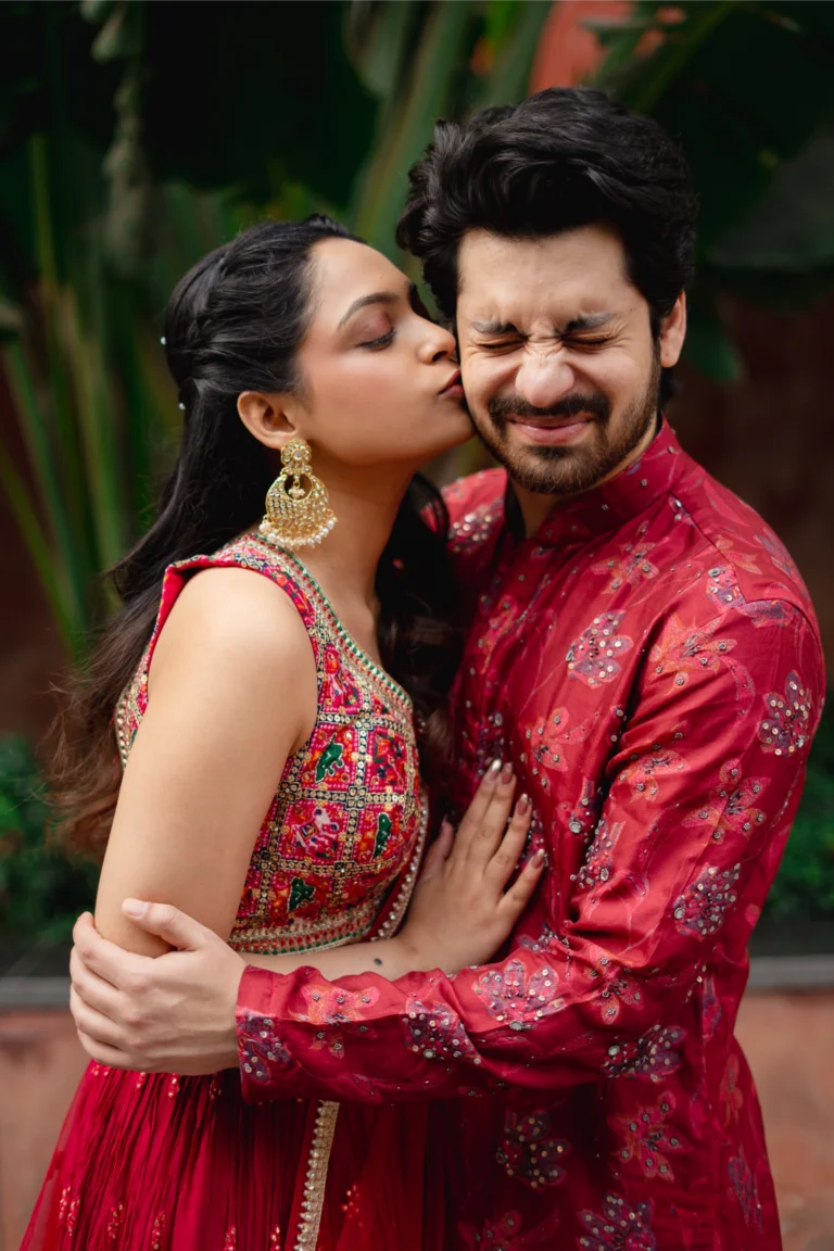 Bride in a red lehenga kissing her laughing groom on the cheek.