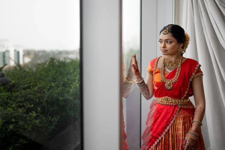 Srija in a red and orange half-saree admiring her mehndi by a window.
