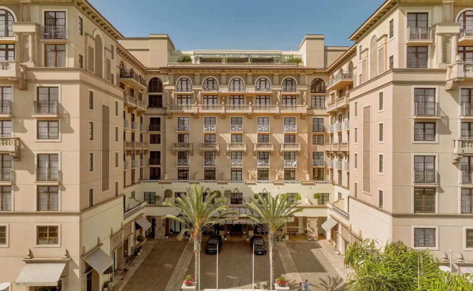The Maybourne Beverly Hills hotel entrance with a cobblestone driveway, two palm trees and cream Mediterranean-style facade with arched balconies.